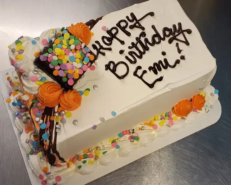 Rectangular cake with white frosting, a brownie with rainbow sprinkles, orange icing roses, and the words Happy Birthday in brown icing