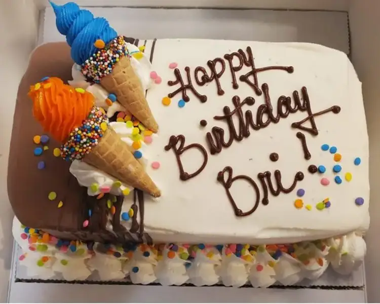 Ice cream cake with white icing, the text Happy Birthday Bri in brown, and two ice cream cones with blue and orange frosting