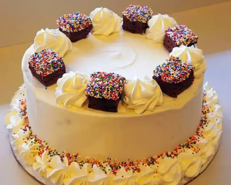 Round ice cream cake with white frosting, brownie squares with sprinkles around the border on top