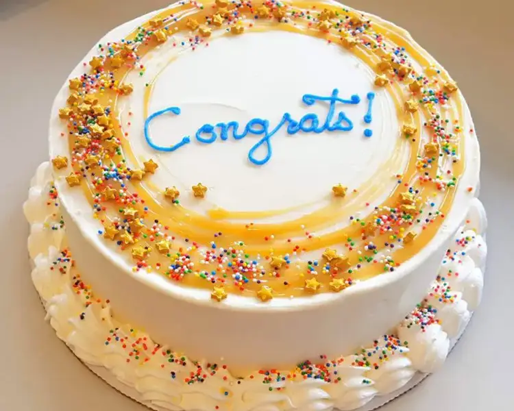 Round cake with white frosting, caramel drizzle and rainbow sprinkles and the word Congrats in blue icing on top