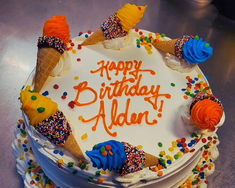Round ice cream cake with white frosting, rainbow sprinkles, and mini cones with different color frosting circling the top